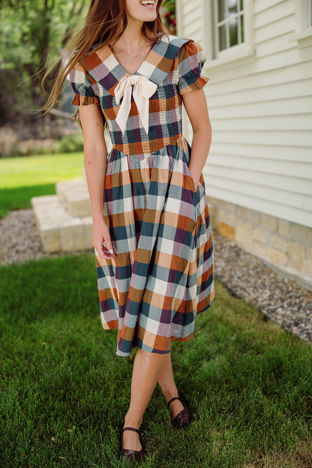 A woman in the Alice Dress in Maize Gingham by Ivy City Co, featuring puff sleeves and a smocked bodice, stands on grass near a white house. She is smiling with her long hair down and has her hands in her pockets.
