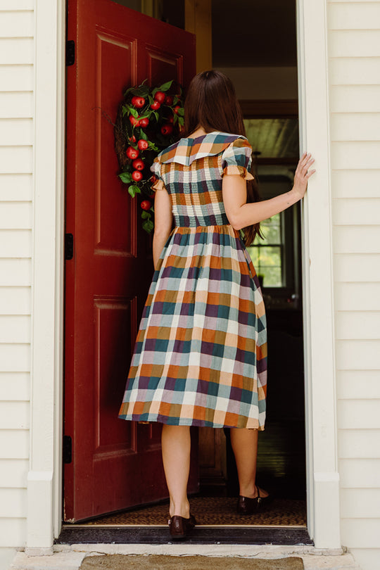 A woman wearing the Ivy City Co Alice Dress in Maize Gingham, featuring puff sleeves and a smocked bodice, enters a house through a red door adorned with a wreath, her back to the camera.
