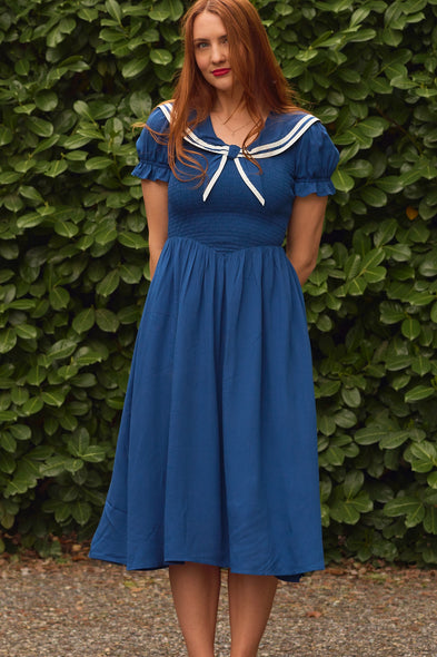 A woman with long red hair stands before green foliage, wearing the Ivy City Co Alice Dress in Navy with a white sailor-style collar and short puffed sleeves, her hands behind her back as she looks at the camera.