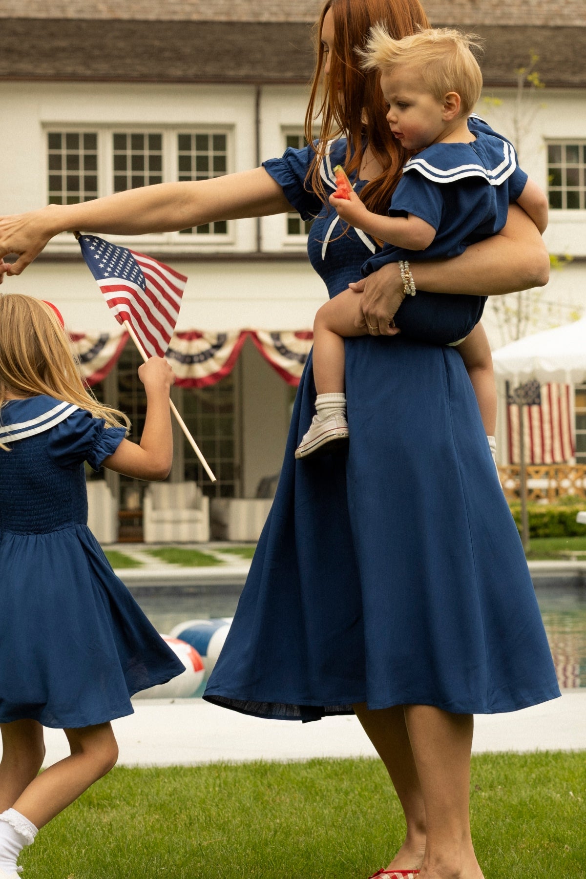A woman wears the Ivy City Co Alice Dress in Sailor Navy as she holds a child in a matching dress; another child, also in Sailor Navy, waves an American flag by the pool with patriotic decorations.