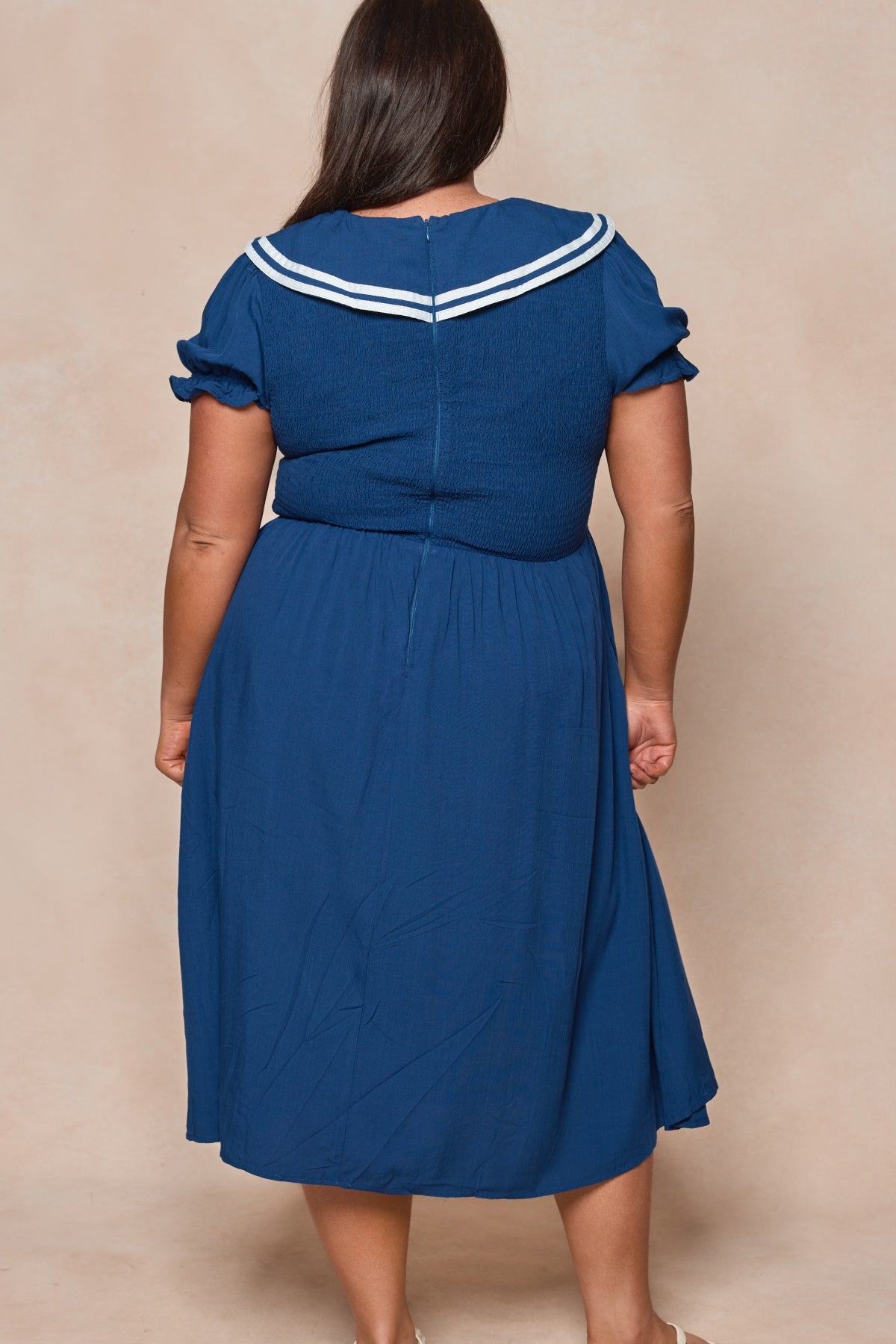 A woman faces away from the camera, wearing the Ivy City Co Alice Dress in Sailor Navy—a summer midi with short sleeves, a white striped sailor collar, fitted bodice, and flared skirt for an effortlessly chic look.
