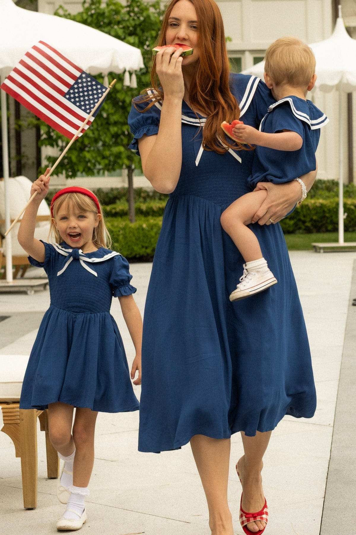 A woman and two children wear Ivy City Co’s Alice Dress in Sailor Navy. The mom holds a toddler and eats watermelon while the older child waves an American flag, showing how this maternity-friendly dress is perfect for sunny summer outings.