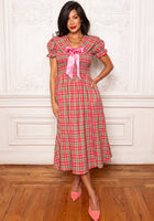 Woman wearing a red and green checkered dress with a pink bow in a room with wooden floor and white walls.