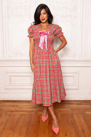 Woman wearing a red and green checkered dress with a pink bow in a room with wooden floor and white walls.