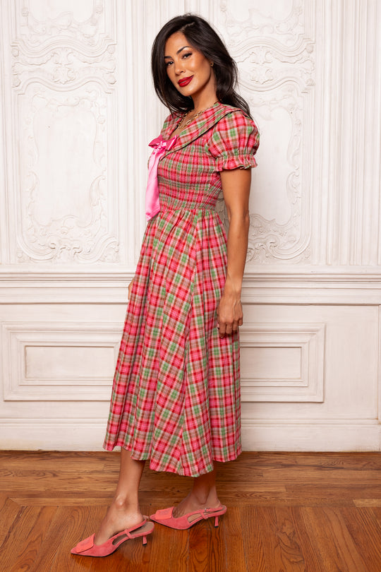 Woman wearing a pink plaid dress with a bow tie in a room with wooden floor and white walls.