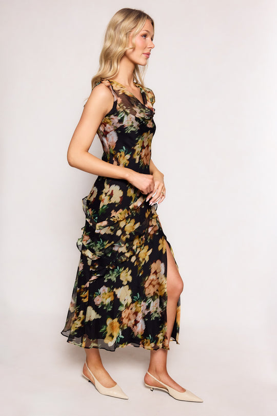 A woman with long blonde hair models the Ivy City Co Alinta Dress in Black Floral, a sleeveless midi dress with a defined waist. She pairs it with beige pointed-toe flats and stands sideways against a plain light background.