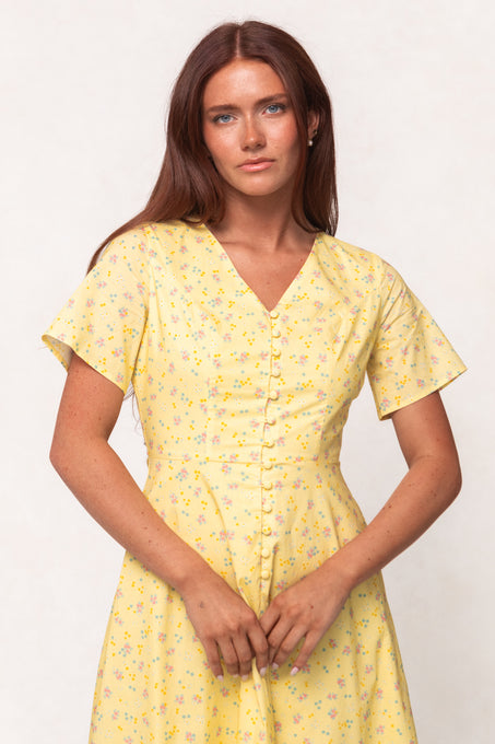 A woman with long brown hair wears the Ivy City Co Allie Dress in Yellow Floral, a nursing-friendly, short-sleeve button-down dress, standing against a plain light background.