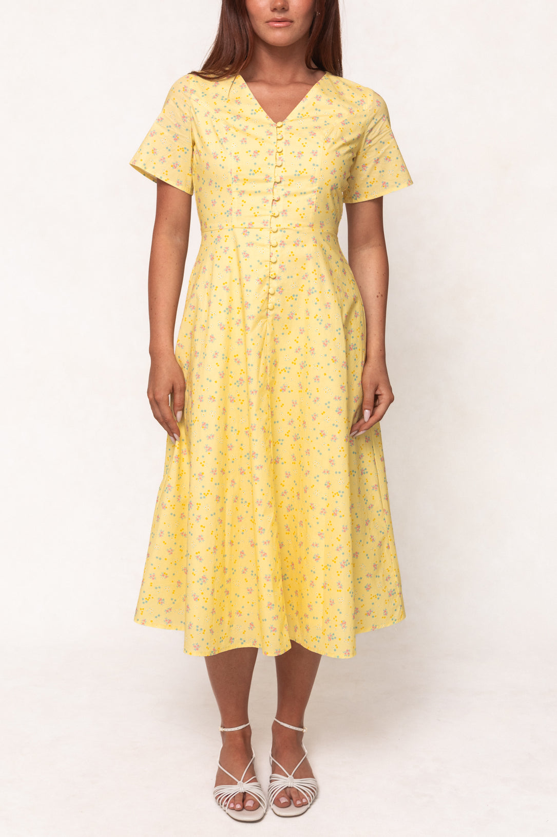 A woman wears the Ivy City Co Allie Dress in Yellow Floral, a nursing-friendly, short-sleeve, button-down dress, paired with white strappy sandals against a plain light background.