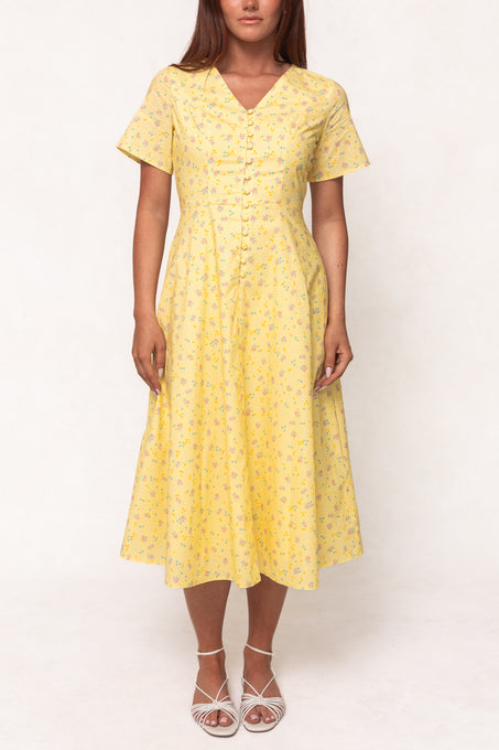 A woman wears the Ivy City Co Allie Dress in Yellow Floral, a nursing-friendly, short-sleeve, button-down dress, paired with white strappy sandals against a plain light background.