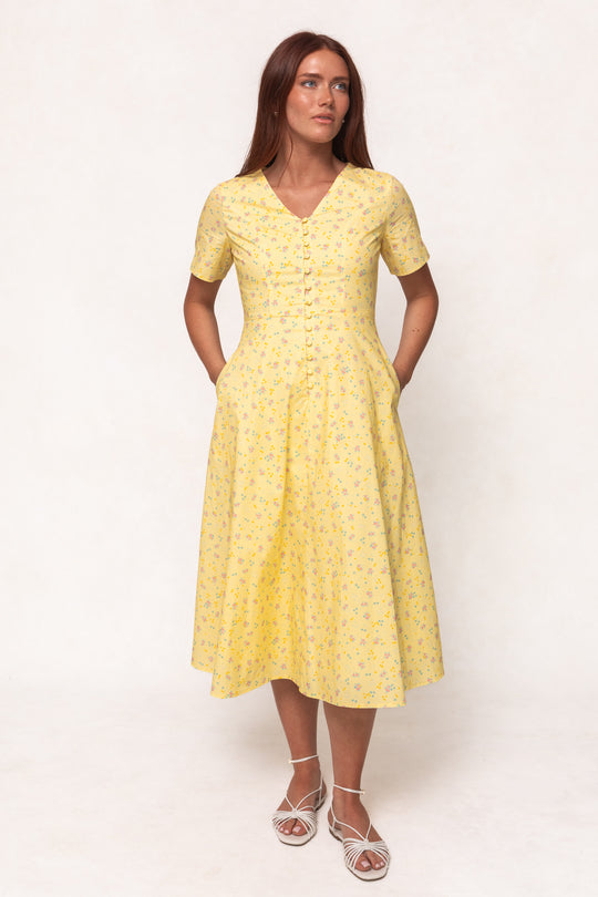 A woman poses against a plain background in the Ivy City Co Allie Dress in Yellow Floral, featuring short sleeves, a button front, and pockets. The mid-length cotton dress is nursing friendly and paired with white strappy sandals.