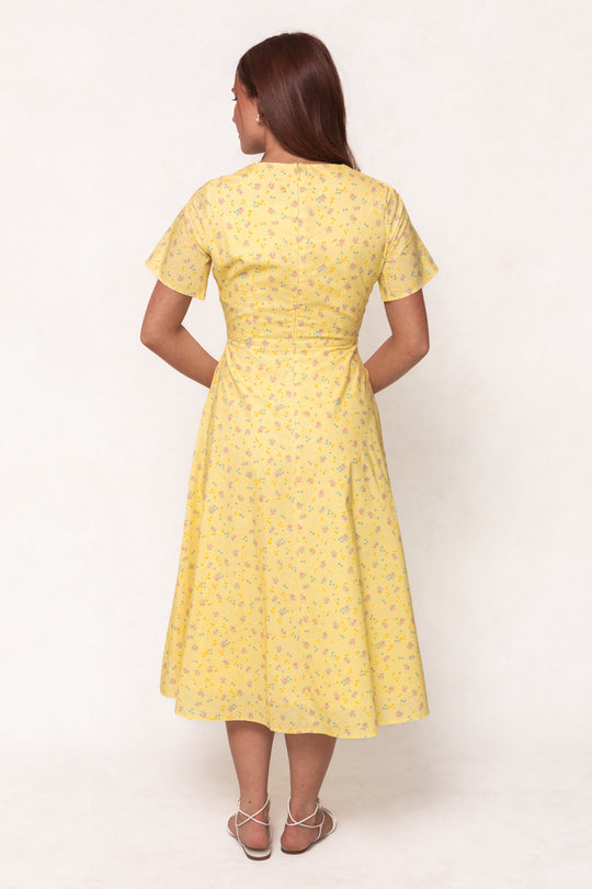 A woman with long brown hair, facing away, wears the Ivy City Co Allie Dress in Yellow Floral—a cotton midi dress with short sleeves and a subtle floral pattern—paired with white strappy sandals against a plain light background.