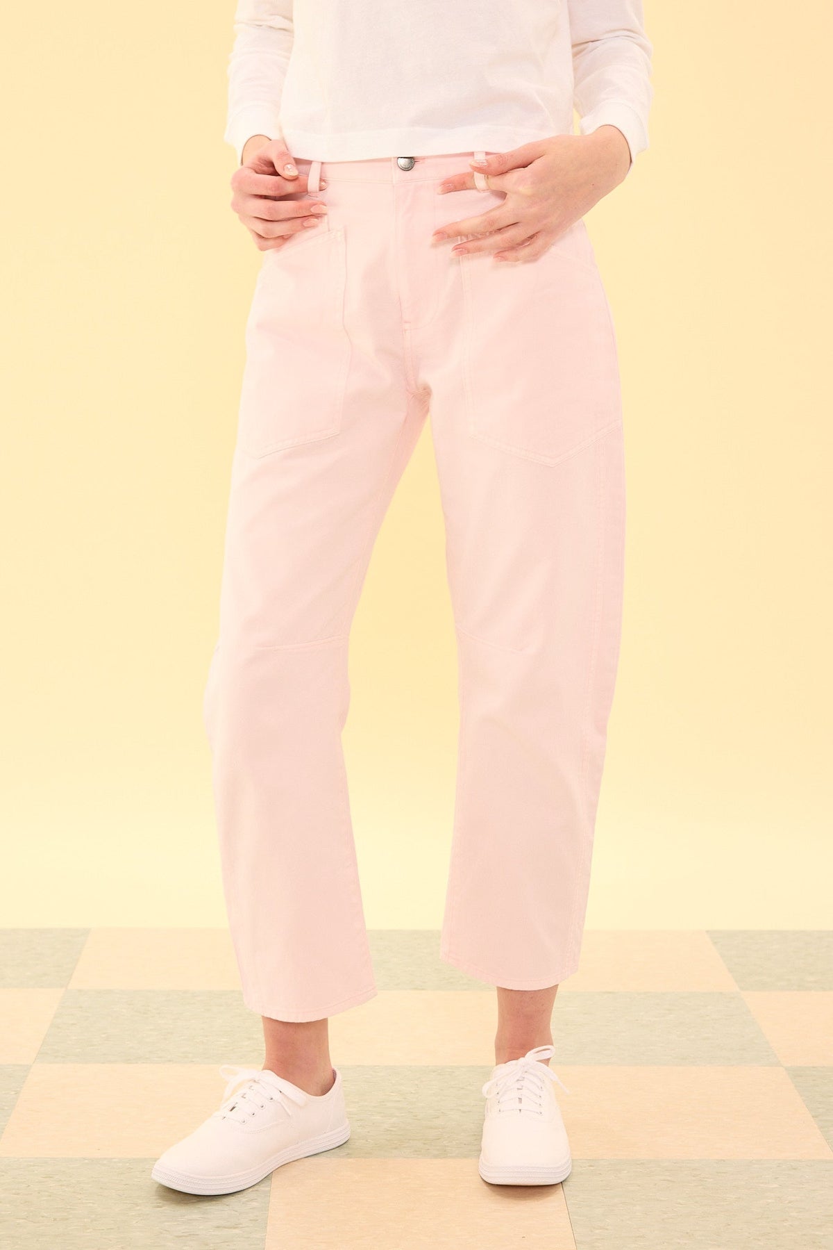 A person wearing the Ivy City Co Amanda Pants in Light Pink, paired with a white long-sleeve shirt and white sneakers, stands on a checkered floor against a pale yellow background. Only the lower half of the body is visible.