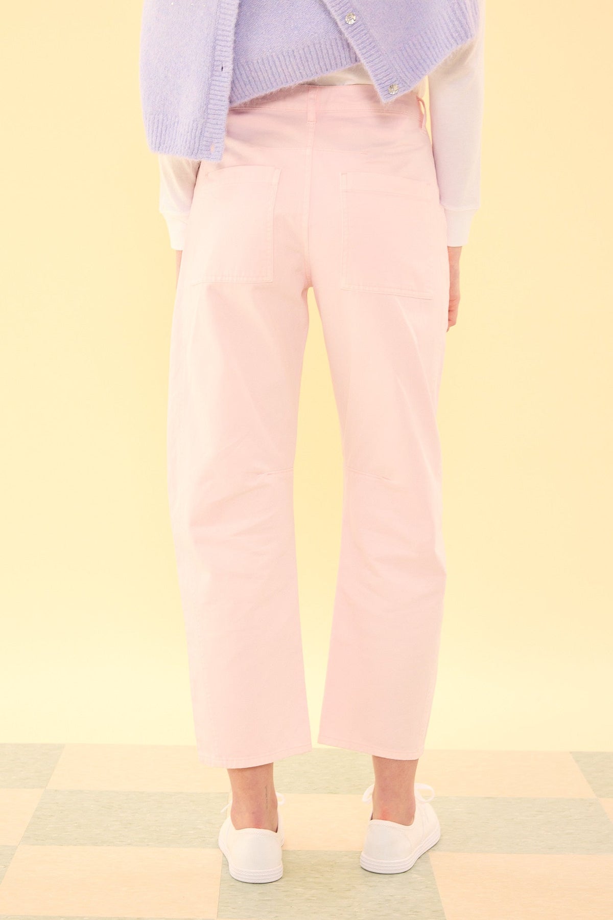 A person stands on a checkered floor, facing away, wearing the Ivy City Co Amanda Pants in Light Pink, paired with white sneakers, a white top, and a lavender sweater draped over their shoulders. The background is a soft yellow.
