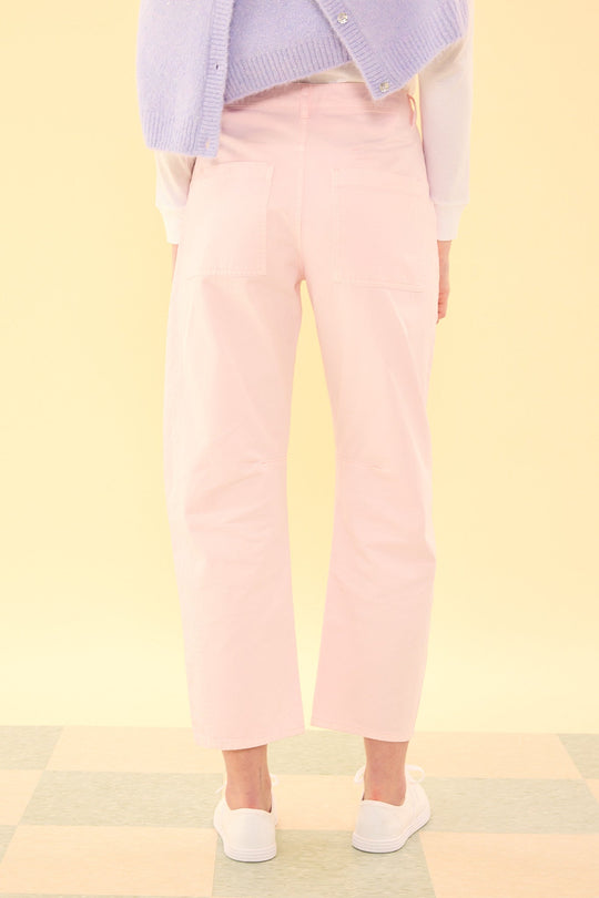 A person stands on a checkered floor, facing away, wearing the Ivy City Co Amanda Pants in Light Pink, paired with white sneakers, a white top, and a lavender sweater draped over their shoulders. The background is a soft yellow.