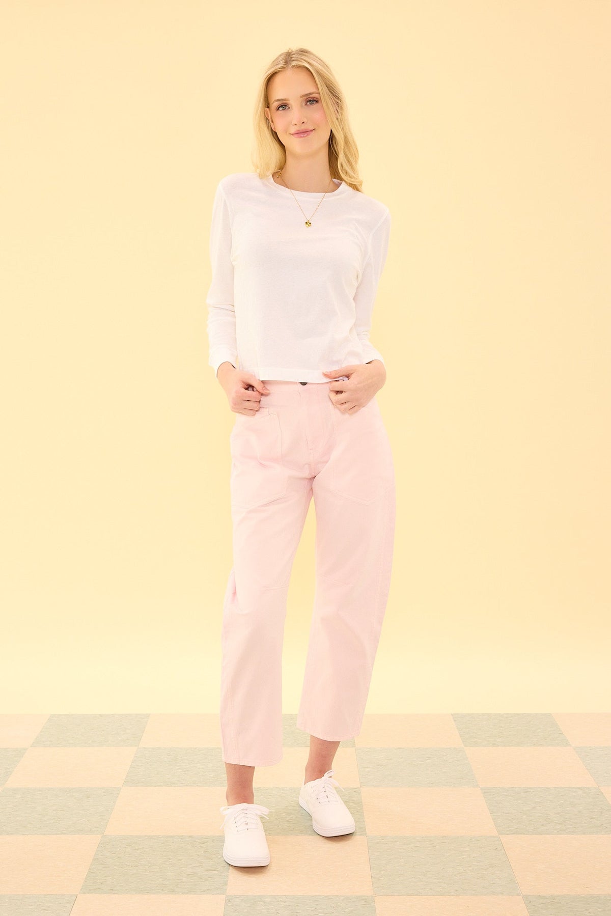 A woman stands on a checkered floor in front of a pale yellow background, wearing the Amanda Pants in Light Pink by Ivy City Co, a white long-sleeve shirt, and white sneakers.