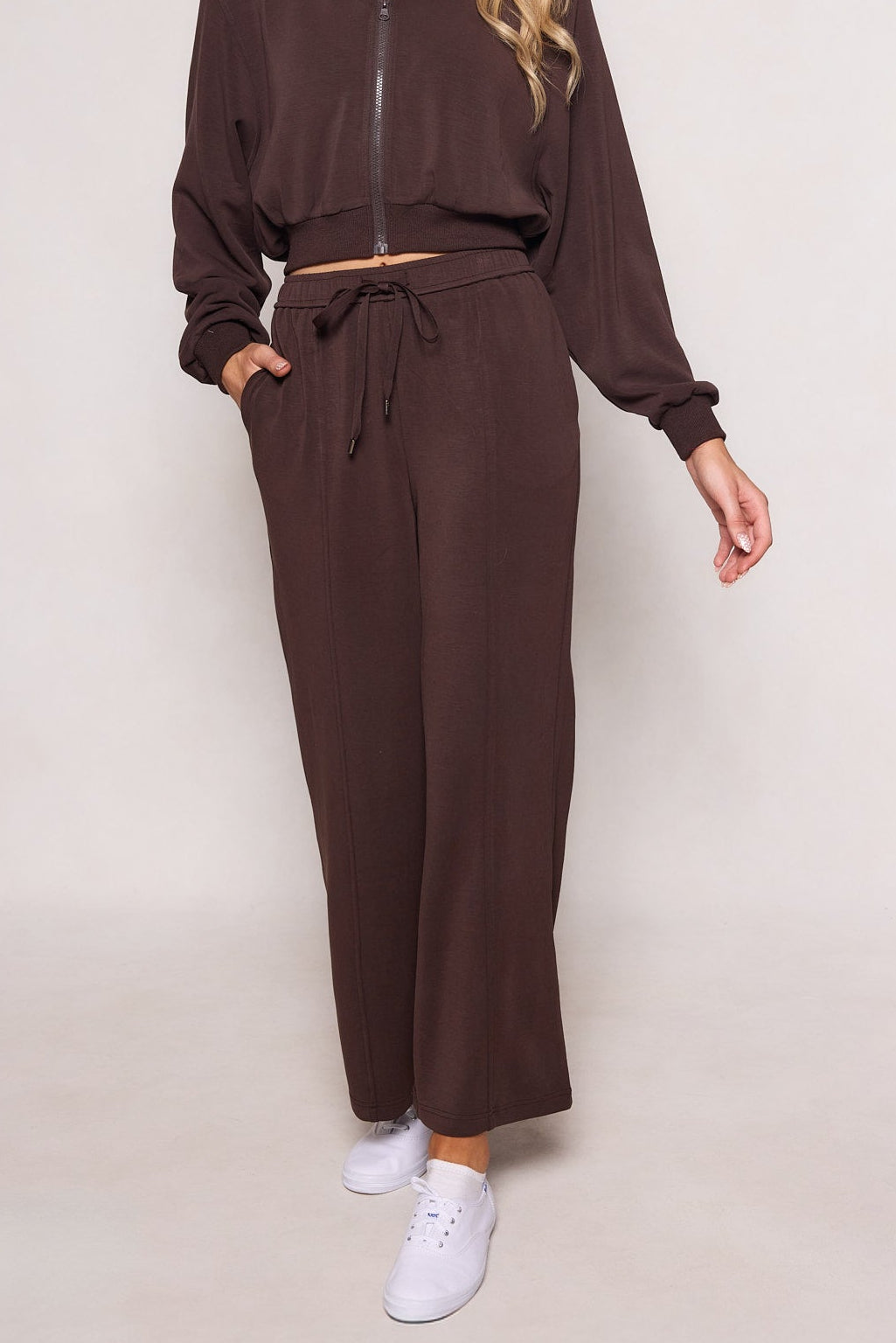 A person wearing the Ivy City Co Amara Sweatpant in Espresso—a high-waisted, relaxed fit pant with an elastic waistband—stands with one hand in a pocket and white sneakers. The head is not visible.