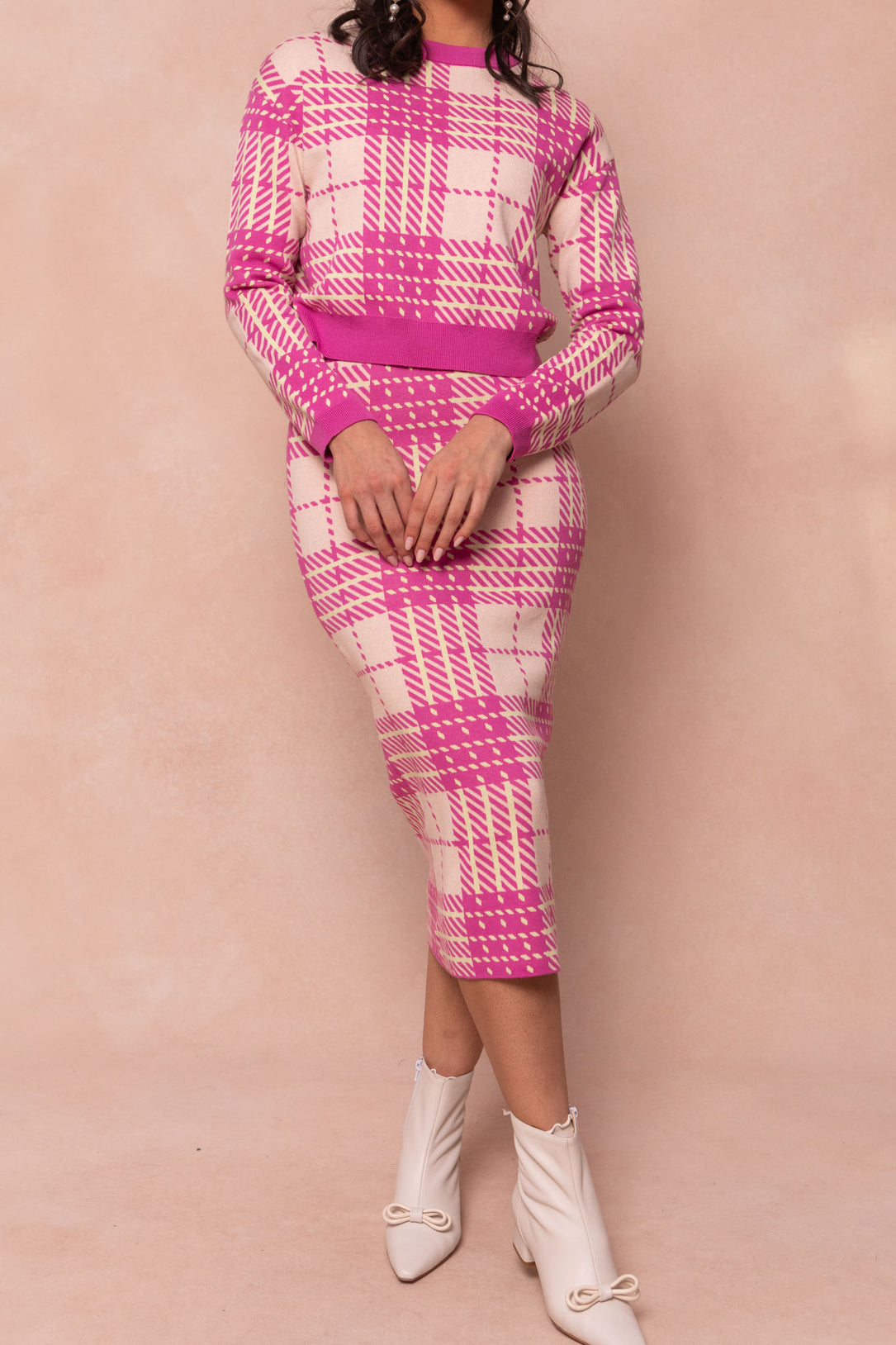 A woman models the Ashley Knit Skirt by Ivy City Co, a pink and cream plaid knit skirt with a matching sweater, styled with white ankle boots with small bows. Her head is not shown, and the background is light beige.