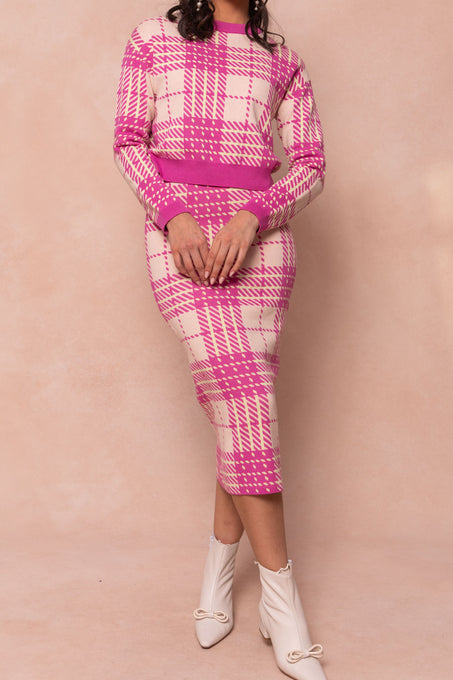 A woman models the Ashley Knit Skirt by Ivy City Co, a pink and cream plaid knit skirt with a matching sweater, styled with white ankle boots with small bows. Her head is not shown, and the background is light beige.