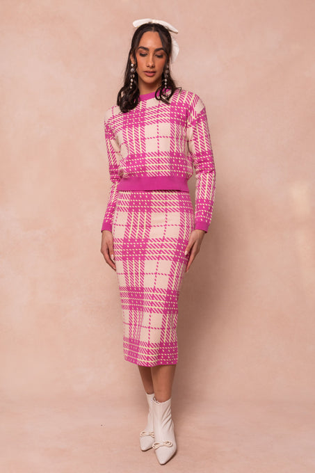 A woman stands against a light beige background, wearing Ivy City Cos Ashley Knit Skirt and matching sweater set in pink and white plaid, styled with white ankle boots, dangling earrings, and a large white hair bow.