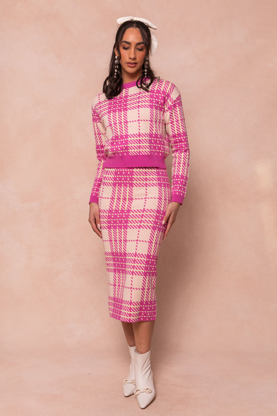 A woman stands against a light beige background, wearing Ivy City Cos Ashley Knit Skirt and matching sweater set in pink and white plaid, styled with white ankle boots, dangling earrings, and a large white hair bow.