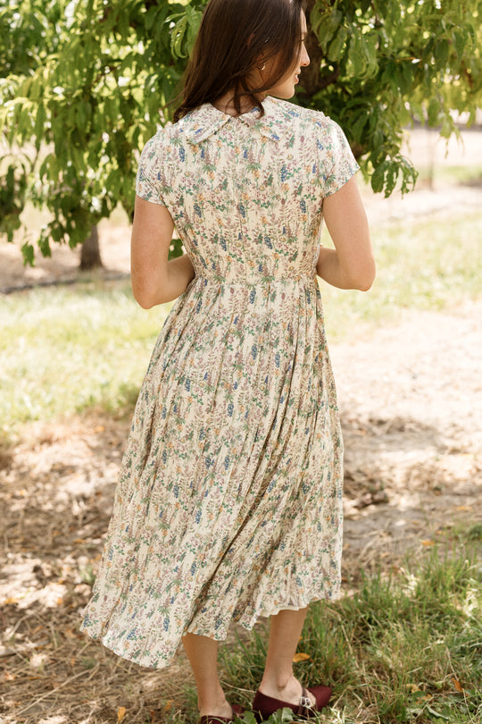 Wearing the Ivy City Co Amber Dress in Autumn Bouquet, a woman stands outdoors beneath a tree, sunlight filtering through the leaves onto her tea-length, nursing-friendly dress and brown shoes as she faces away.