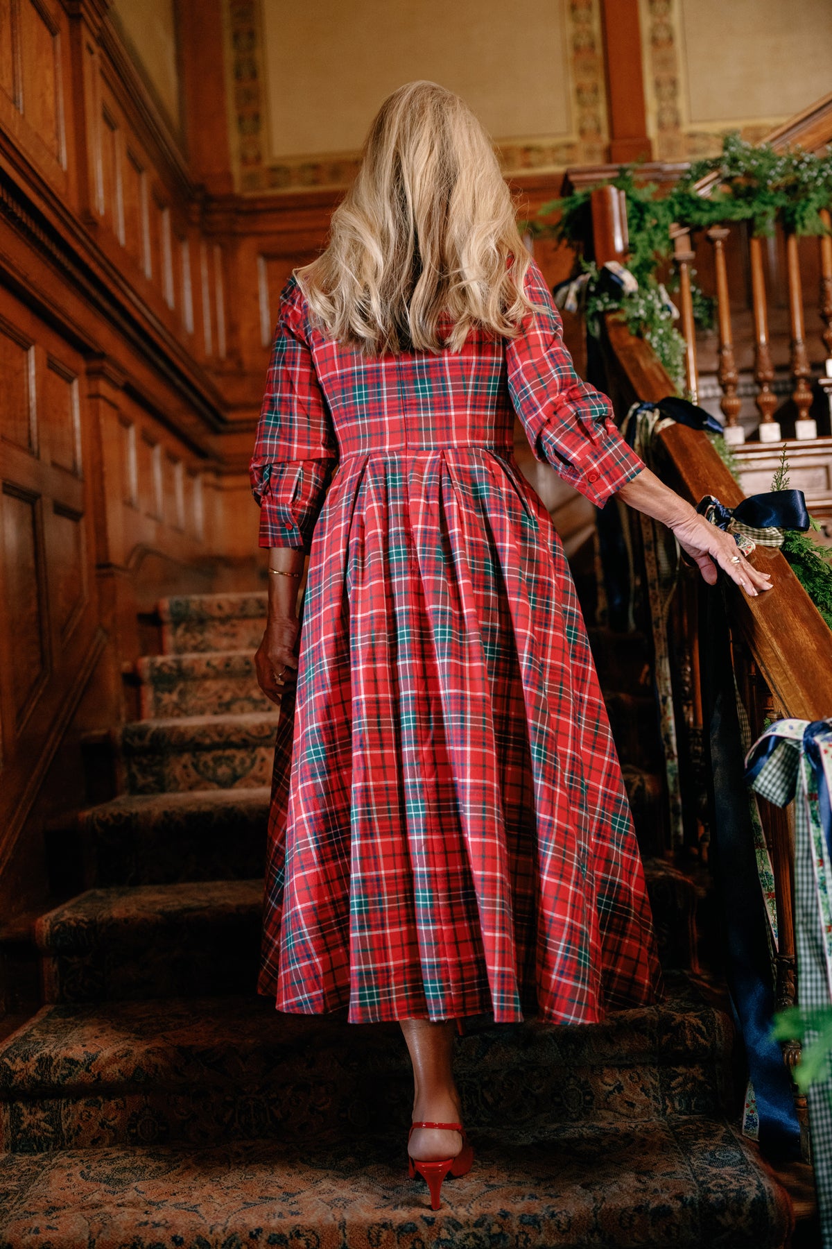 A woman with long blonde hair, dressed in the Ivy City Co Amber 3/4 Sleeve Dress in Christmas Cheer Plaid and red heels, walks up a carpeted staircase in a wood-paneled, festively decorated interior.