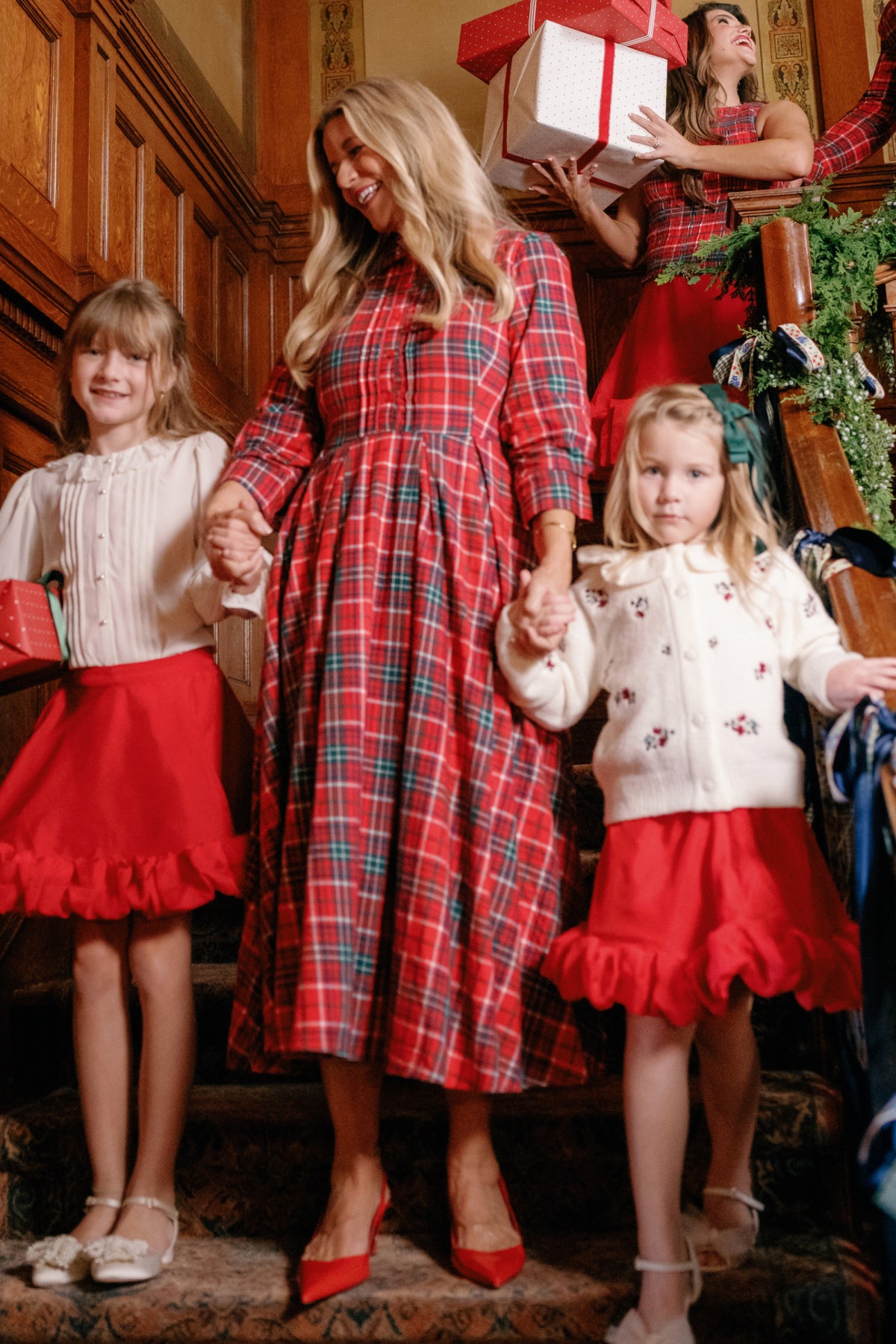 A woman wearing the Ivy City Co Amber 3/4 Sleeve Dress in Christmas Cheer Plaid holds hands with two young girls dressed in white tops and red Christmas skirts, standing on holiday-decorated stairs. In the background, another woman in red carries presents.