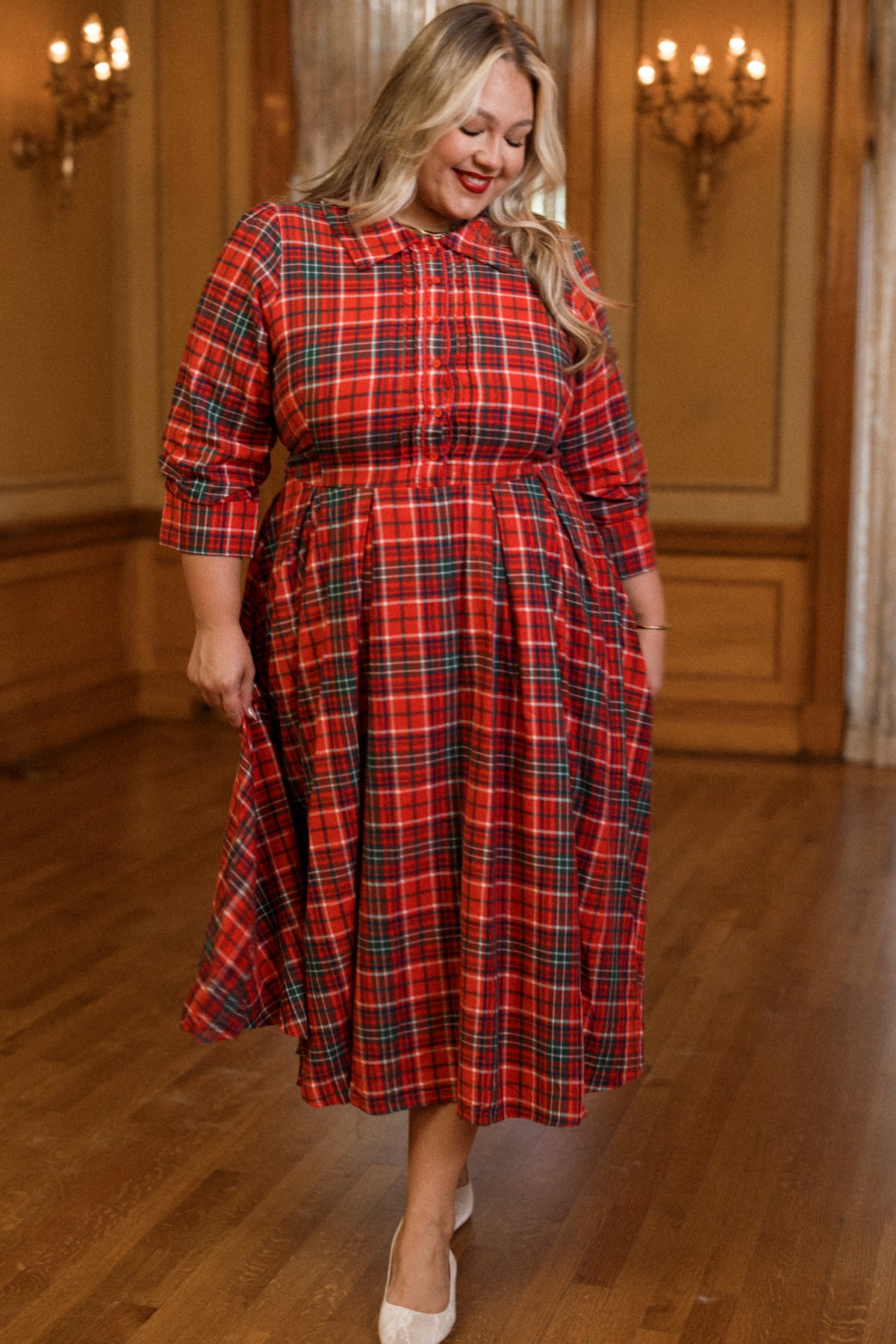 A woman wearing the Ivy City Co Amber 3/4 Sleeve Dress in Christmas Cheer Plaid smiles and looks down while standing in an elegant room with wooden floors and chandeliers.