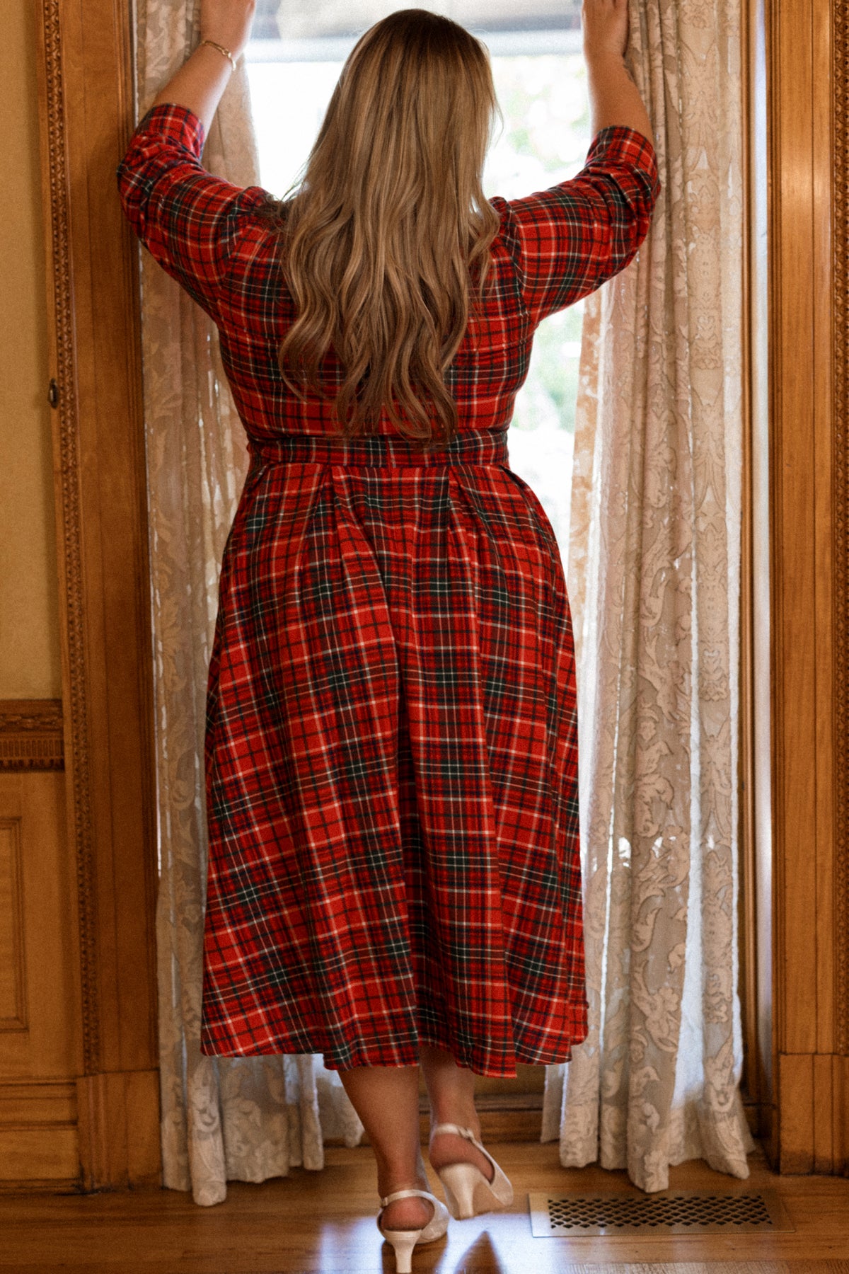 Wearing the Ivy City Co Amber 3/4 Sleeve Dress in Christmas Cheer Plaid, a woman with long blonde hair stands by a window, holding back lace curtains as she gazes outside.
