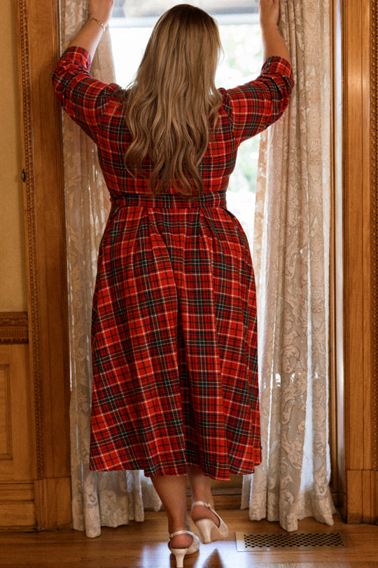 Wearing the Ivy City Co Amber 3/4 Sleeve Dress in Christmas Cheer Plaid, a woman with long blonde hair stands by a window, holding back lace curtains as she gazes outside.