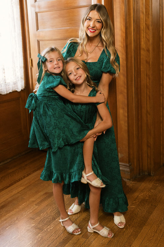 Three individuals—a woman and two young girls—stand together on a wooden floor in a warmly lit room accented with wood paneling. All three are wearing matching Ivy City Co “Amira Dress in Emerald Satin” designs, paired with white sandals. The girls smile brightly; one embraces the other while being held up.