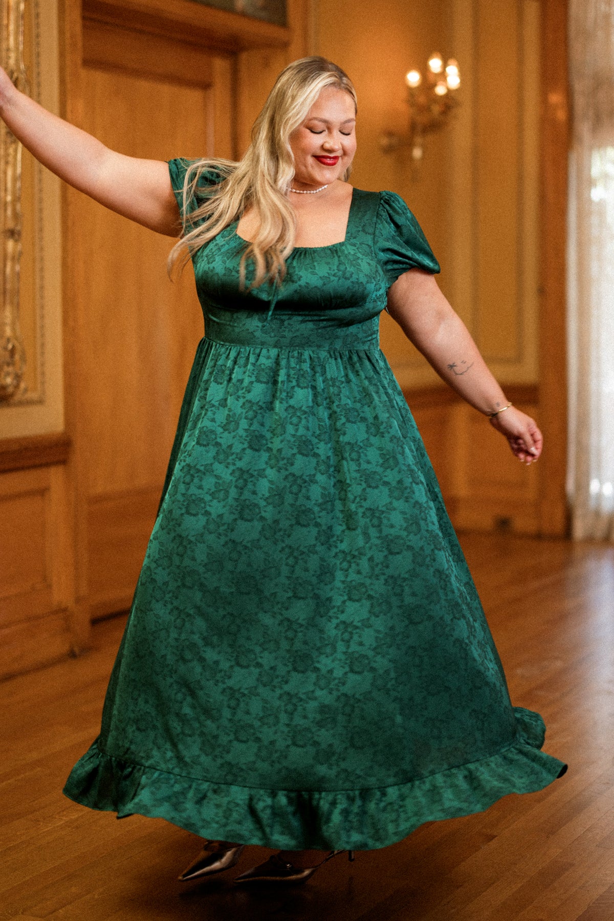 Wearing the Amira Dress in Emerald Satin by Ivy City Co, a woman twirls with a smile and closed eyes on a wooden floor, showcasing its short puff sleeves and elegant floral pattern.