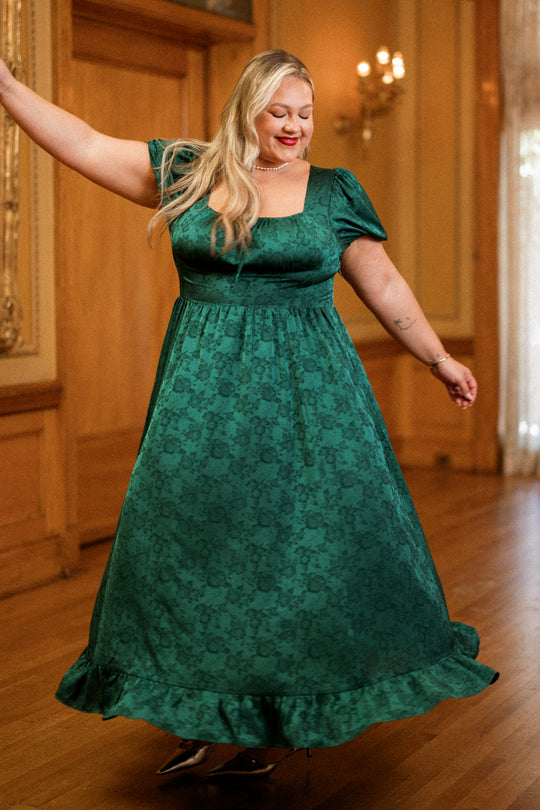 Wearing the Amira Dress in Emerald Satin by Ivy City Co, a woman twirls with a smile and closed eyes on a wooden floor, showcasing its short puff sleeves and elegant floral pattern.