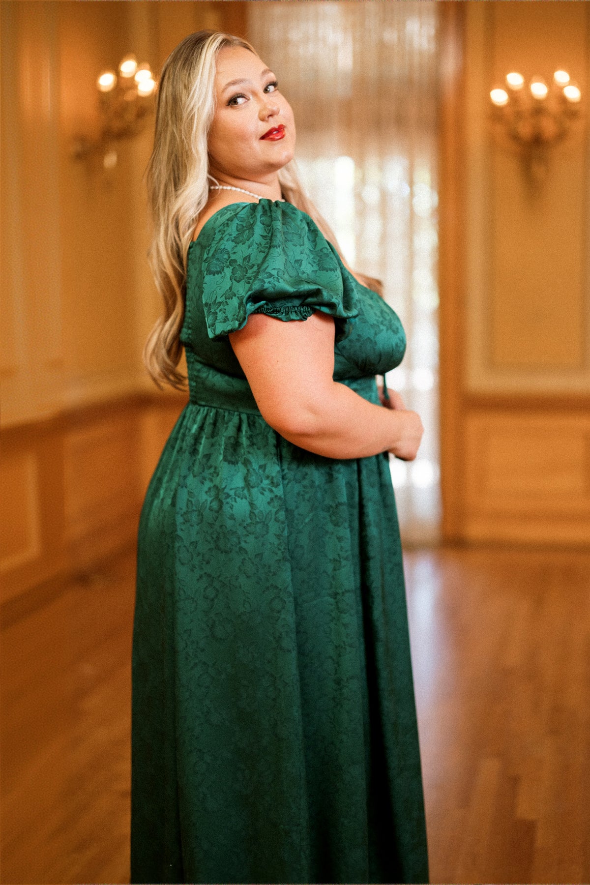 A woman wearing the Amira Dress in Emerald Satin by Ivy City Co stands in an elegant room with wooden floors and chandeliers, looking over her shoulder and smiling confidently at the camera.