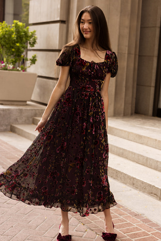 A woman in a flowy, dark floral Amira Dress in Violet Dusk Burnout by Ivy City Co with puffed sleeves walks along a brick path, smiling. This nursing-friendly maxi dress with pockets offers both style and practicality to her look. In the background, stone stairs and a large planter overflowing with green plants add to the charming setting.