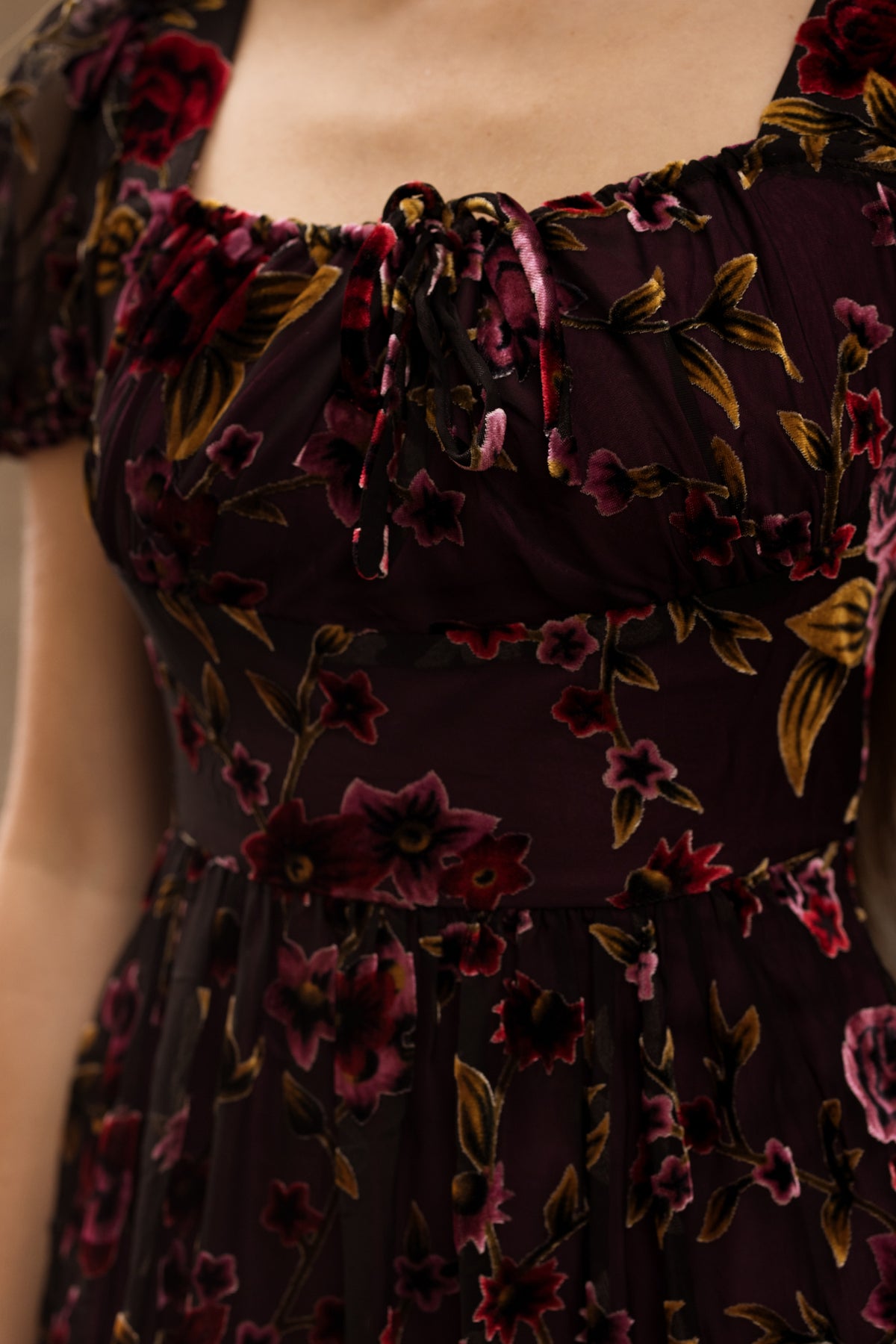 Close-up of a woman’s torso wearing the Amira Dress in Violet Dusk Burnout by Ivy City Co—a dark, sheer maxi dress with pockets and a floral pattern in shades of red, pink, and gold. The dress features short sleeves, a gathered tied neckline, and is nursing friendly for added convenience.