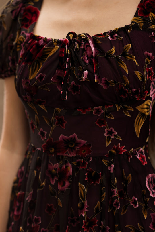 Close-up of a woman’s torso wearing the Amira Dress in Violet Dusk Burnout by Ivy City Co—a dark, sheer maxi dress with pockets and a floral pattern in shades of red, pink, and gold. The dress features short sleeves, a gathered tied neckline, and is nursing friendly for added convenience.