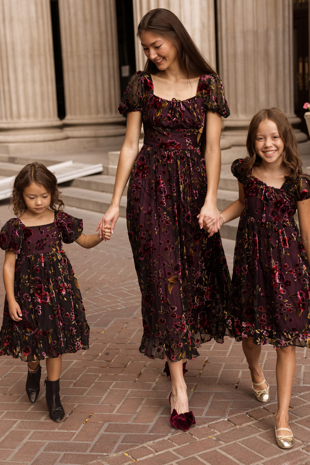 A woman and two young girls, all wearing matching Ivy City Co Amira Dresses in Violet Dusk Burnout, walk hand in hand on a brick path lined with tall columns. The dark floral maxi dresses with pockets are both stylish and nursing friendly, making them an ideal choice for family outings.