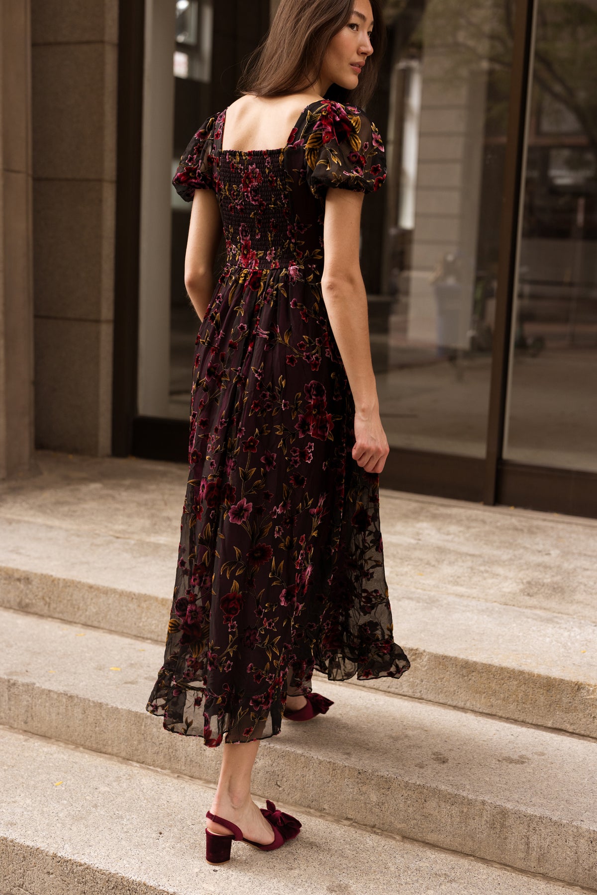 A woman in the Ivy City Co Amira Dress in Violet Dusk Burnout—a nursing-friendly, dark floral midi silhouette with puff sleeves—pairs it with burgundy heels as she walks up stone steps outside a building with large windows.