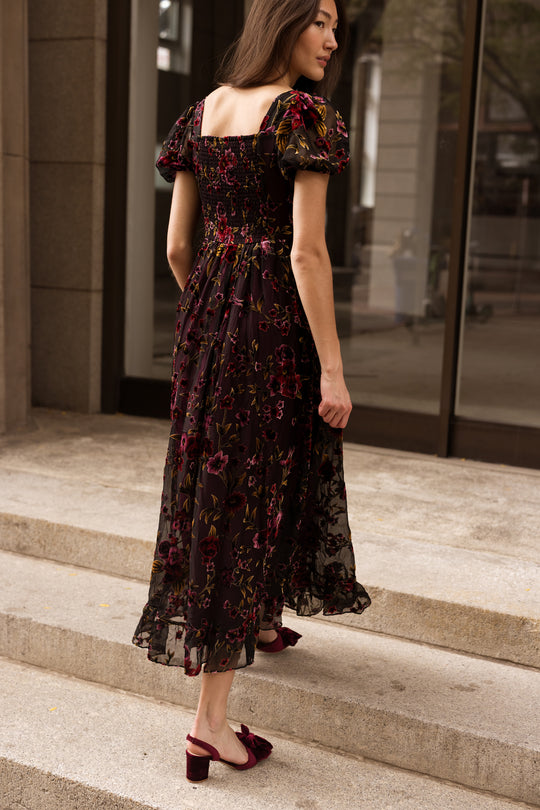 A woman in the Ivy City Co Amira Dress in Violet Dusk Burnout—a nursing-friendly, dark floral midi silhouette with puff sleeves—pairs it with burgundy heels as she walks up stone steps outside a building with large windows.