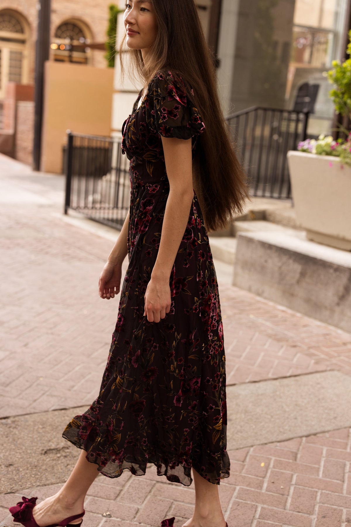 A woman with long brown hair walks outdoors wearing the Amira Dress in Violet Dusk Burnout by Ivy City Co, a dark floral maxi dress with pockets, paired with maroon heels. She is smiling, with buildings and plants in the background.