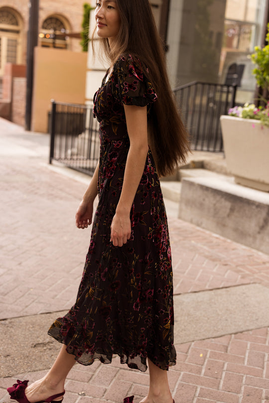 A woman with long brown hair walks outdoors wearing the Amira Dress in Violet Dusk Burnout by Ivy City Co, a dark floral maxi dress with pockets, paired with maroon heels. She is smiling, with buildings and plants in the background.