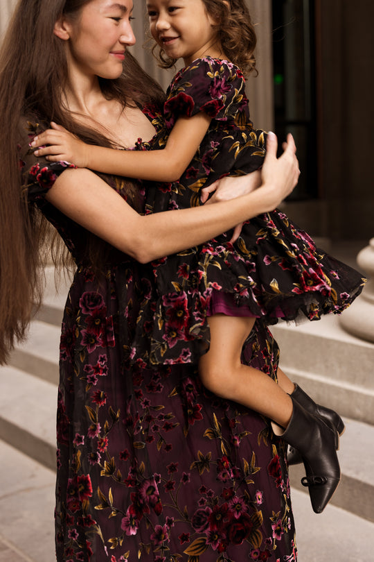A woman holds a smiling young girl outside, both dressed in matching floral outfits. The woman wears the nursing-friendly Amira Dress in Violet Dusk Burnout by Ivy City Co, and the girl sports black boots. They share a joyful moment together near stone steps.