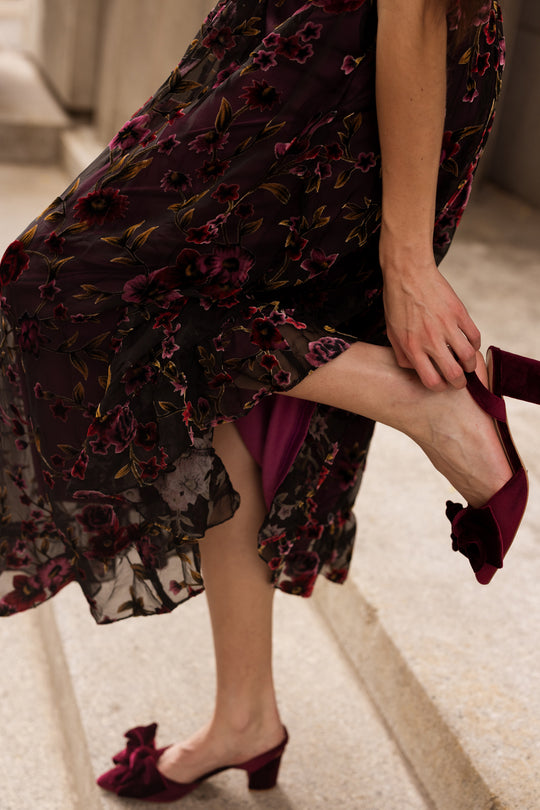 A person wearing the Ivy City Co Amira Dress in Violet Dusk Burnout adjusts a burgundy heeled shoe with a bow while standing on stone steps.