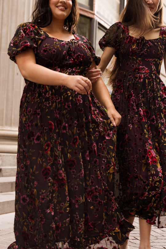 Two women in matching Ivy City Co Amira Dresses in Violet Dusk Burnout walk side by side outdoors on stone steps, smiling and holding their skirts, with a columned building in the background.