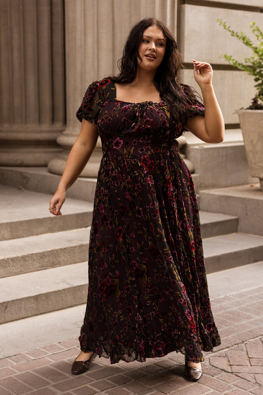 A woman wearing the Ivy City Co Amira Dress in Violet Dusk Burnout stands outdoors by stone steps and columns, smiling and lifting her hand. The nursing-friendly maxi dress with pockets pairs beautifully with her long dark hair and flat shoes.