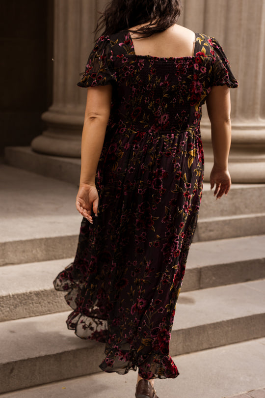 A person wearing the Amira Dress in Violet Dusk Burnout by Ivy City Co walks up stone steps outdoors, framed by pillars in the background. The photo, taken from behind, highlights the nursing-friendly design of this dark floral maxi dress with pockets and showcases its graceful movement.