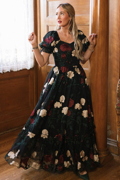 A woman stands indoors by wooden columns and a white lace-curtained window, looking to the side with her hands raised near her shoulders. She wears the Amora Dress in Obsidian Rose Embroidery from Ivy City Co, a long black gown adorned with red and white floral details, featuring elegant puff sleeves and a sweetheart neckline.