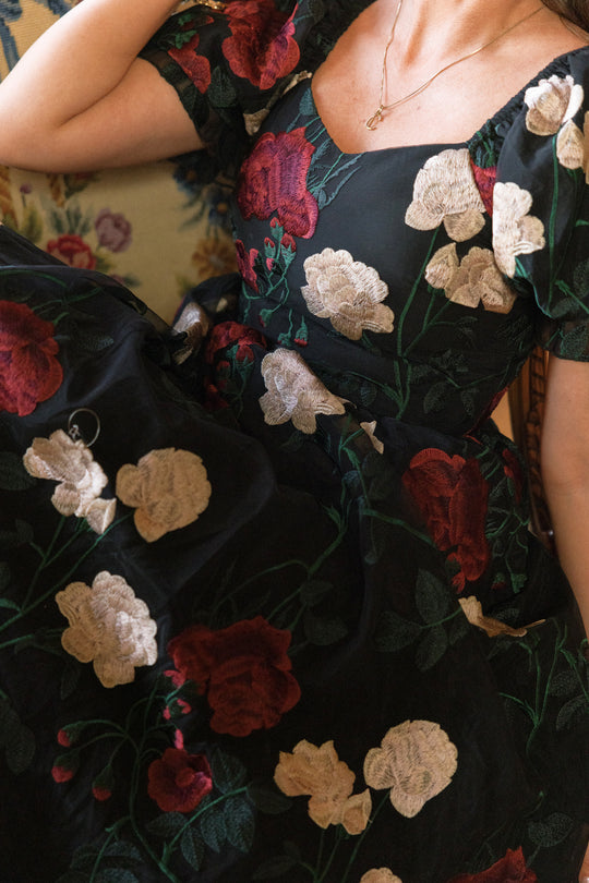 A person wears the Amora Dress in Obsidian Rose Embroidery by Ivy City Co, featuring a black puff-sleeve silhouette with a square neckline and striking, oversized red and cream floral embroidery. They accessorize with a simple gold necklace and are seated on a floral-patterned chair.