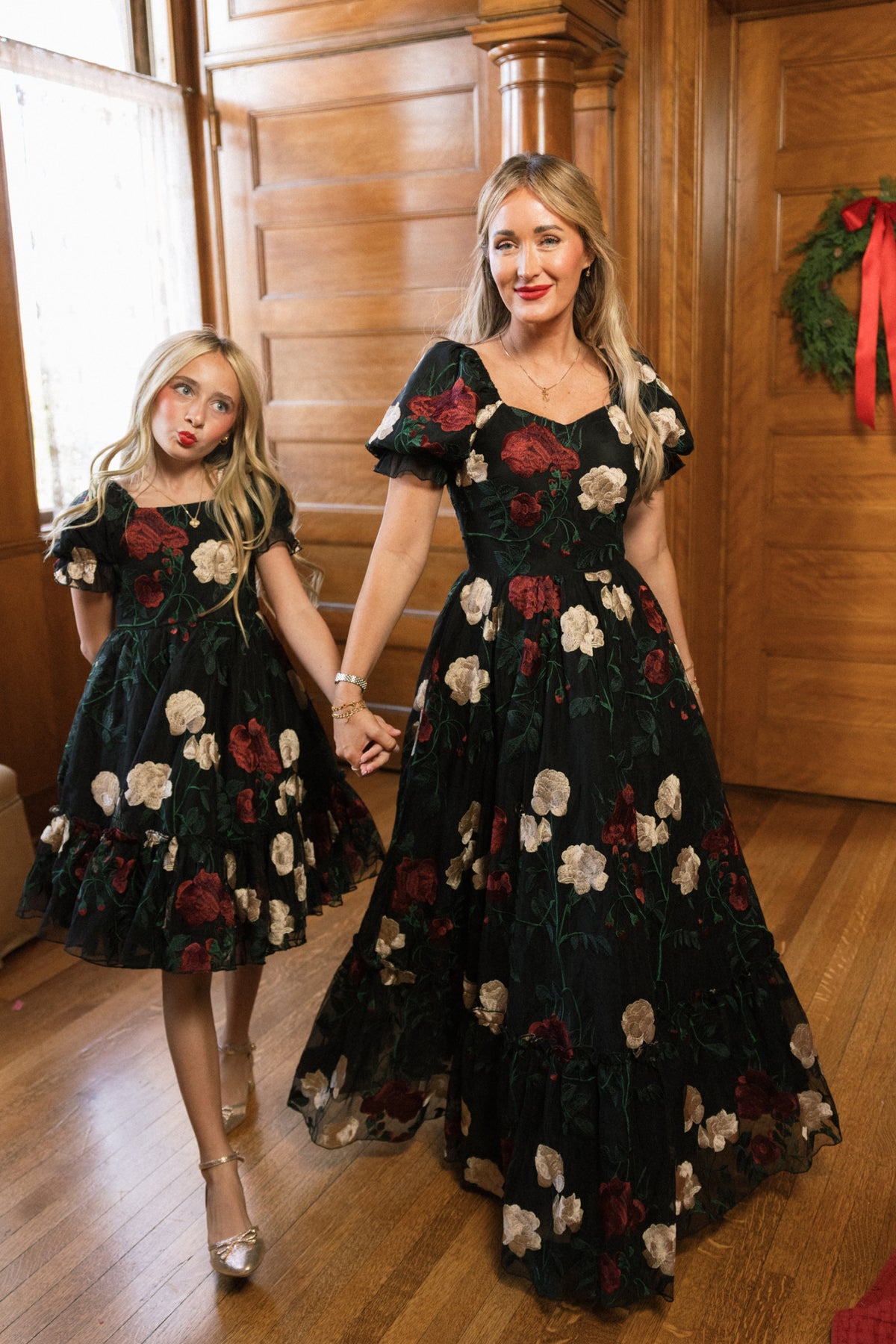 A woman and a girl hold hands, both wearing matching Ivy City Co Amora Dresses in Obsidian Rose Embroidery—black tulle ball gowns featuring red and white floral patterns with puff sleeve sweetheart necklines—as they stand in a wood-paneled room adorned with holiday decor.